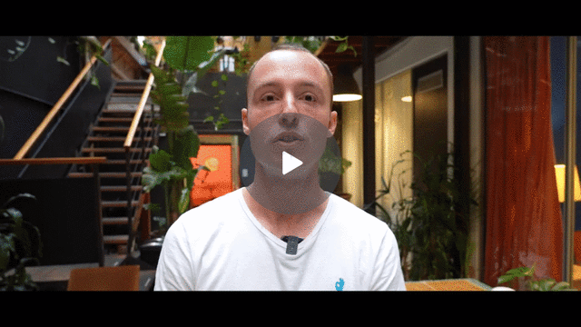 A bald man in a white shirt is speaking indoors in this video.