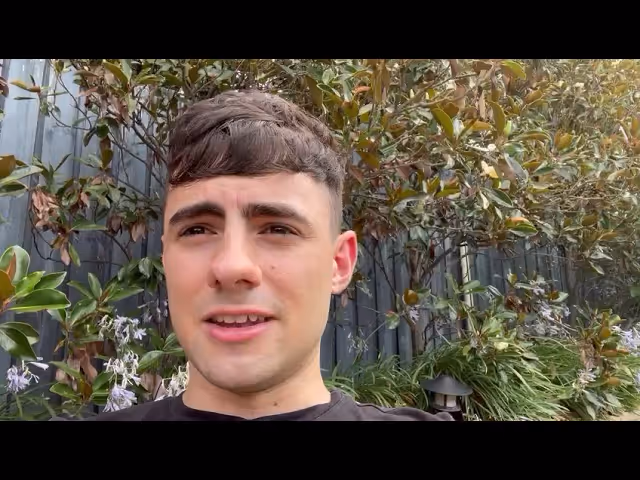 Young man with short dark hair speaking outdoors with leafy plants and a fence in the background.